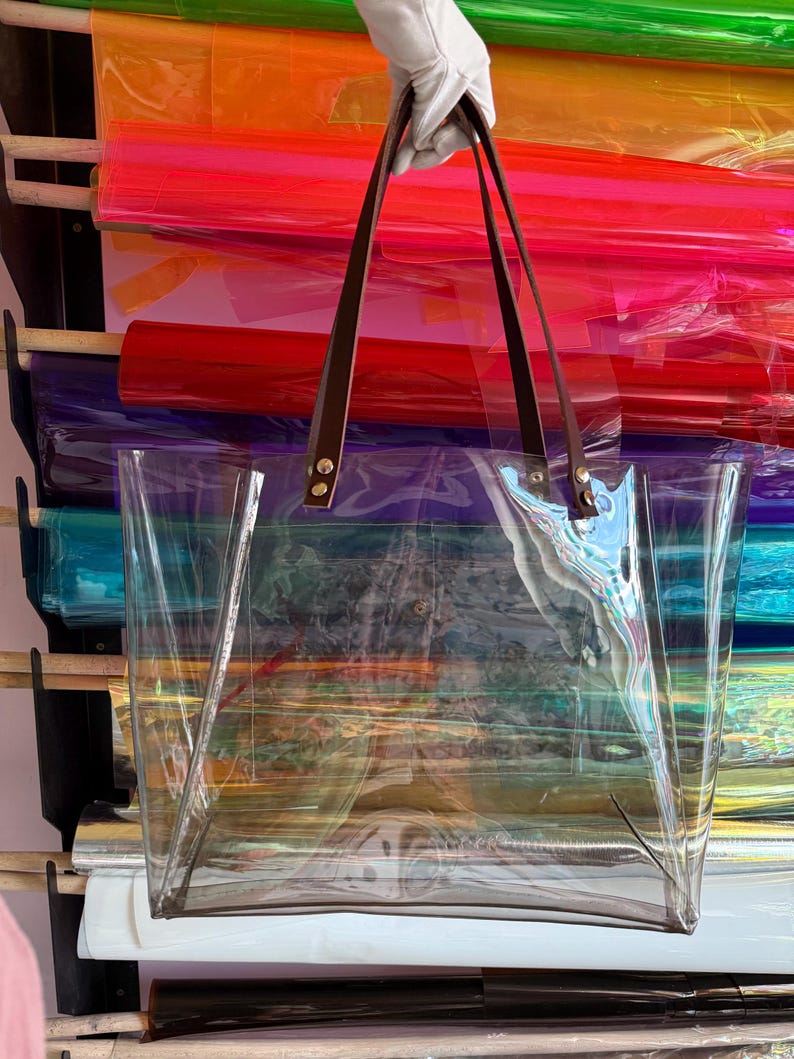 Puede incluir: Un bolso tote transparente e incoloro con dos correas de hombro de cuero marr&oacute;n oscuro sujetas con remaches de metal plateado. El bolso tiene un dise&ntilde;o rectangular de apertura superior con un compartimento principal y base plana.