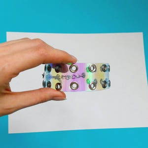 May include: Iridescent choker bracelet with silver-tone grommets and a delicate chain. The bracelet has a holographic effect, displaying a range of colors. The accessory is held against a white background with a blue backdrop.