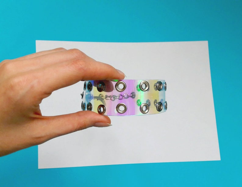 May include: Iridescent choker bracelet with silver-tone grommets and a delicate chain. The bracelet has a holographic effect, displaying a range of colors. The accessory is held against a white background with a blue backdrop.