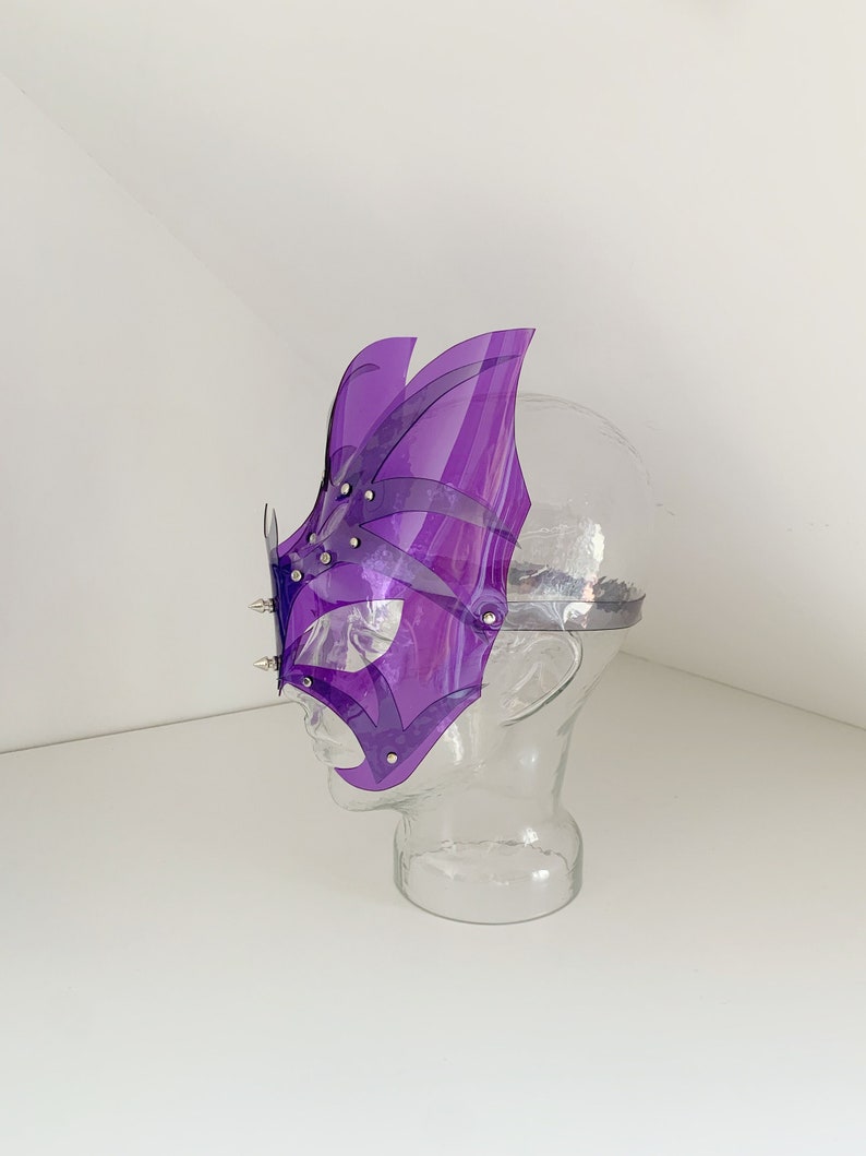 Op de afbeelding: Paarse plastic maskerbalmasker met een geometrisch ontwerp en zilveren studs. Het masker bevindt zich op een mannequinhoofd.