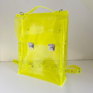 May include: A neon yellow, transparent backpack with a top handle and adjustable shoulder strap. The bag features silver-colored metal clasps and hardware. The design is rectangular with rounded corners and a flap closure.