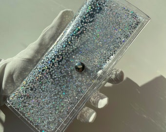 Könnte beinhalten: Eine transparente, rechteckige Hülle, gefüllt mit silbernem Glitzer in Sternform. Die Hülle hat einen silbernen Druckknopfverschluss und wird von einer behandschuhten Hand gehalten. Die Hülle ist für kleine Gegenstände konzipiert.