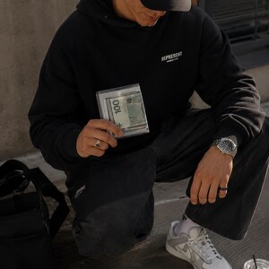 May include: A black hooded sweatshirt with the text REPRESENT OWNERS CLUB printed in white on the chest, paired with black trousers, a black baseball cap, a silver wristwatch, and a clear plastic wallet containing a 100 Polish Zloty banknote.