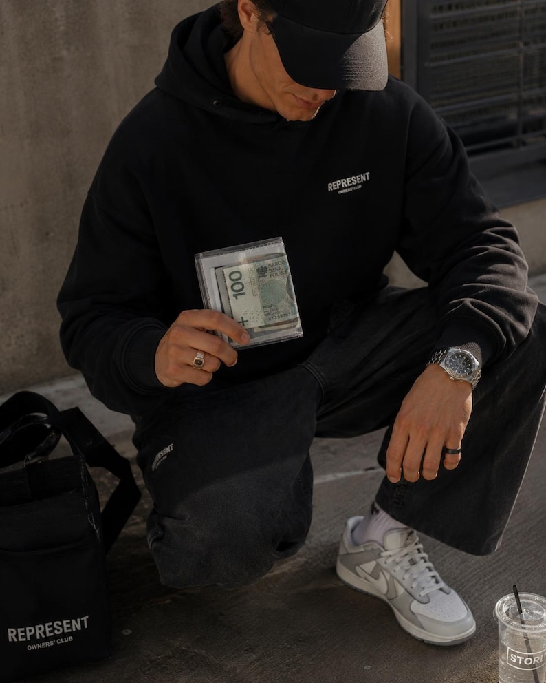 May include: A black hooded sweatshirt with the text REPRESENT OWNERS CLUB printed in white on the chest, paired with black trousers, a black baseball cap, a silver wristwatch, and a clear plastic wallet containing a 100 Polish Zloty banknote.