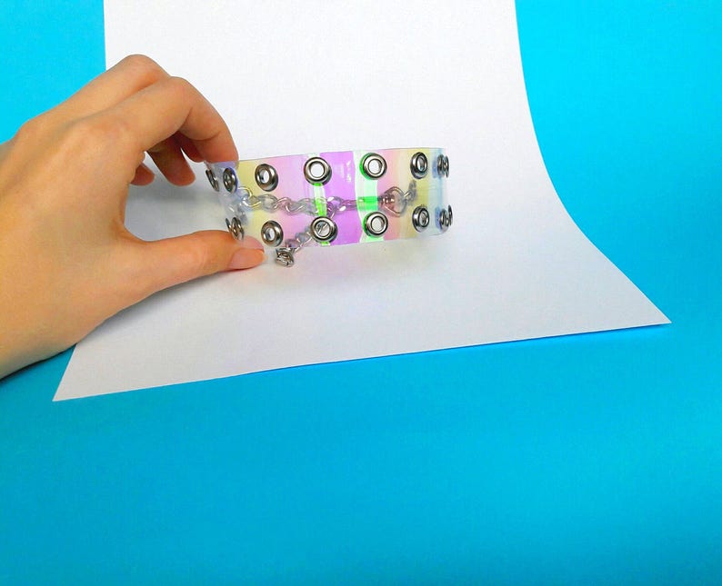 May include: A clear, iridescent choker with silver-tone metal grommets and a delicate chain closure. The choker features a rainbow-colored stripe design and is held by a hand against a white and blue background.
