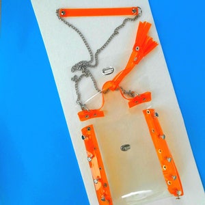 May include: A clear, rectangular bag with bright orange accents and silver chain detailing. The bag features orange straps, rivets, and a chain strap. The design includes a small, silver clasp and a decorative orange tassel.