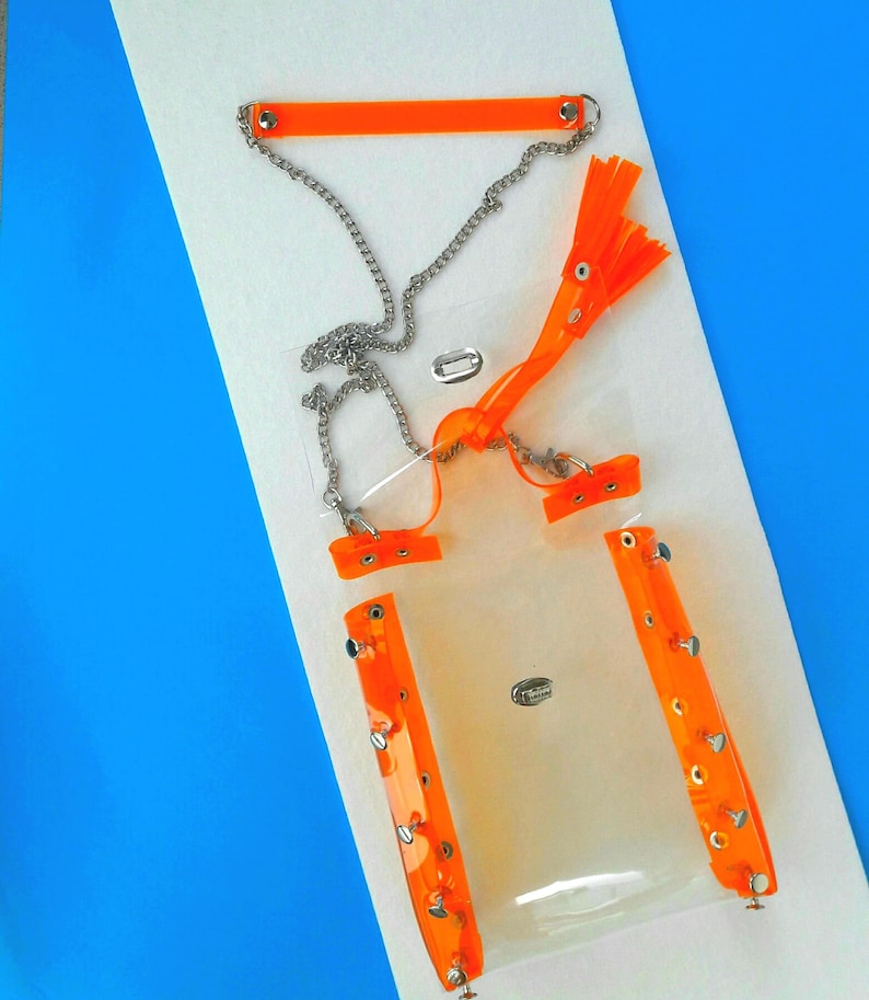 May include: A clear, rectangular bag with bright orange accents and silver chain detailing. The bag features orange straps, rivets, and a chain strap. The design includes a small, silver clasp and a decorative orange tassel.