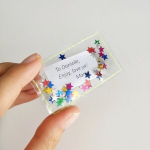 May include: A clear plastic bag with a yellow stitched border contains colorful star-shaped confetti and a white note that reads "To Danielle, Enjoy, love ya! Mary".