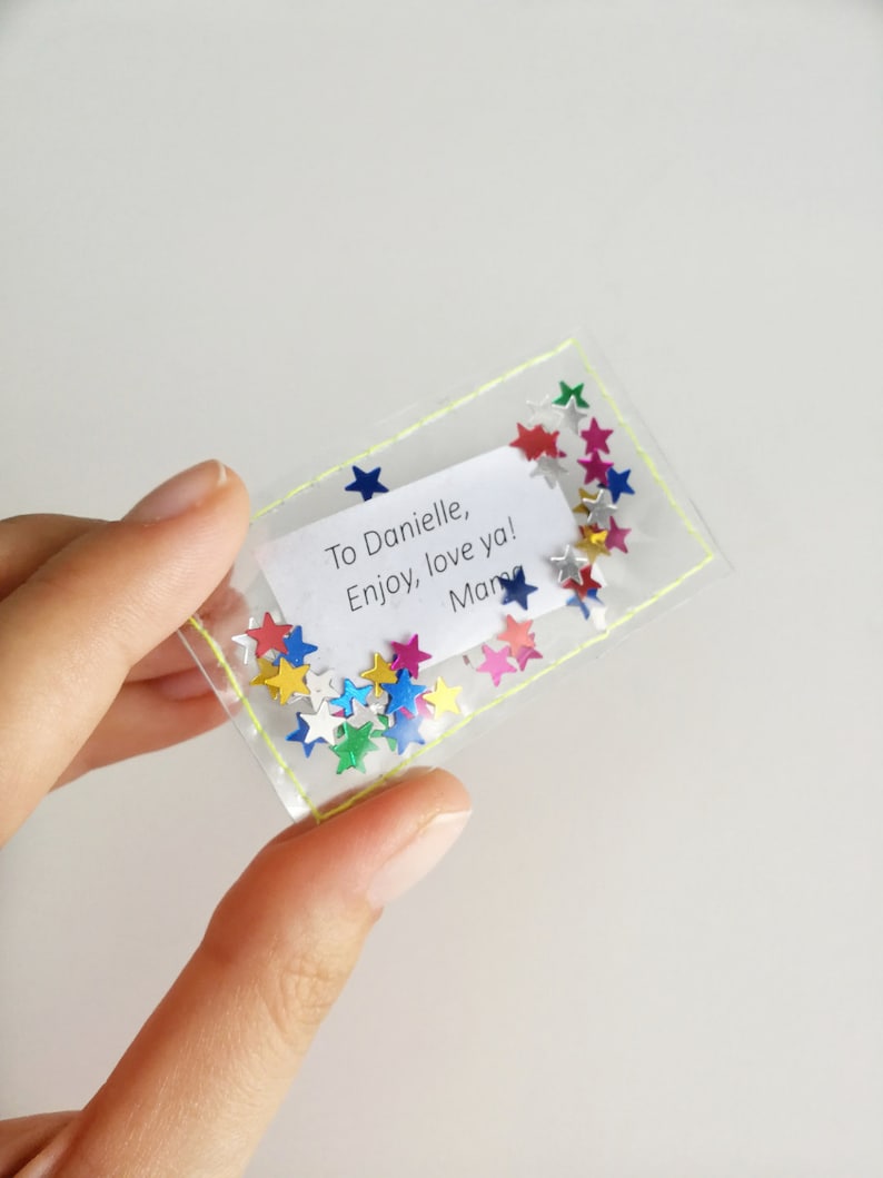 May include: A clear plastic bag with a yellow stitched border contains colorful star-shaped confetti and a white note that reads "To Danielle, Enjoy, love ya! Mary".