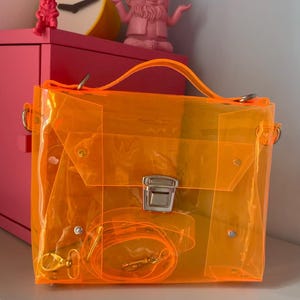 May include: A transparent orange handbag with a top handle and a detachable shoulder strap. The bag has a rectangular shape with a silver clasp and hardware. The bag is sitting on a white surface.