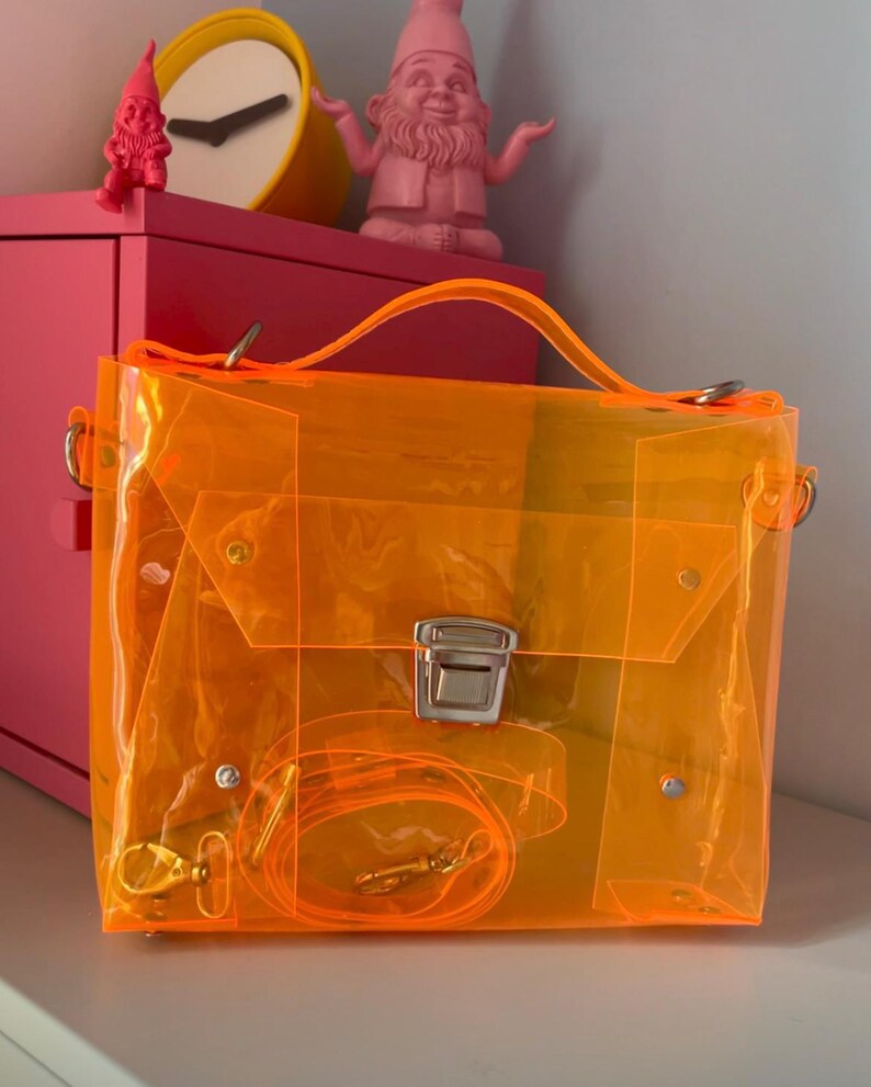 May include: A transparent orange handbag with a top handle and a detachable shoulder strap. The bag has a rectangular shape with a silver clasp and hardware. The bag is sitting on a white surface.