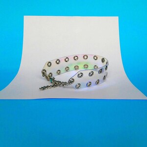 May include: A clear, iridescent choker necklace with silver-colored grommets and a chain closure. The choker is made of a flexible, transparent material that reflects a rainbow of colors. The necklace is displayed on a white surface against a blue background.