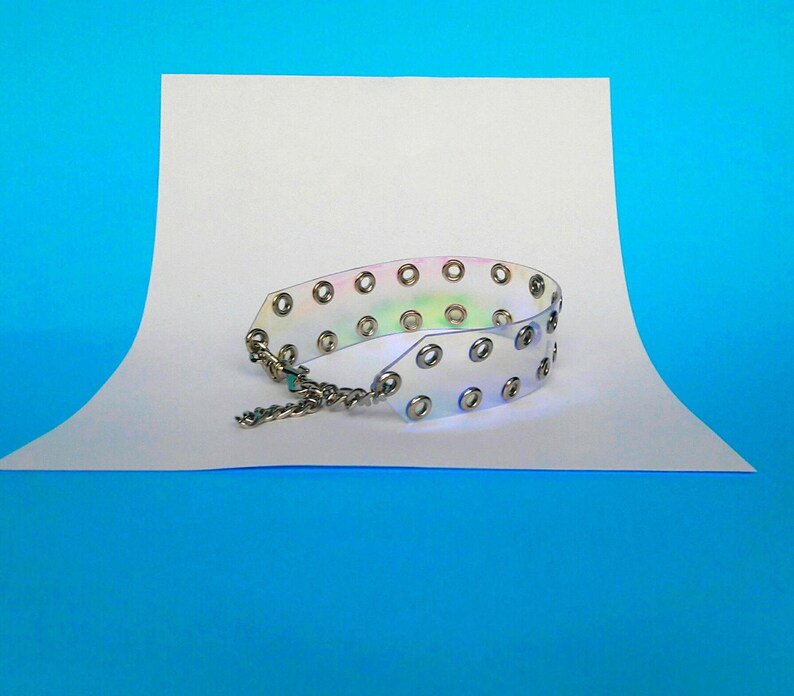 May include: A clear, iridescent choker necklace with silver-colored grommets and a chain closure. The choker is made of a flexible, transparent material that reflects a rainbow of colors. The necklace is displayed on a white surface against a blue background.