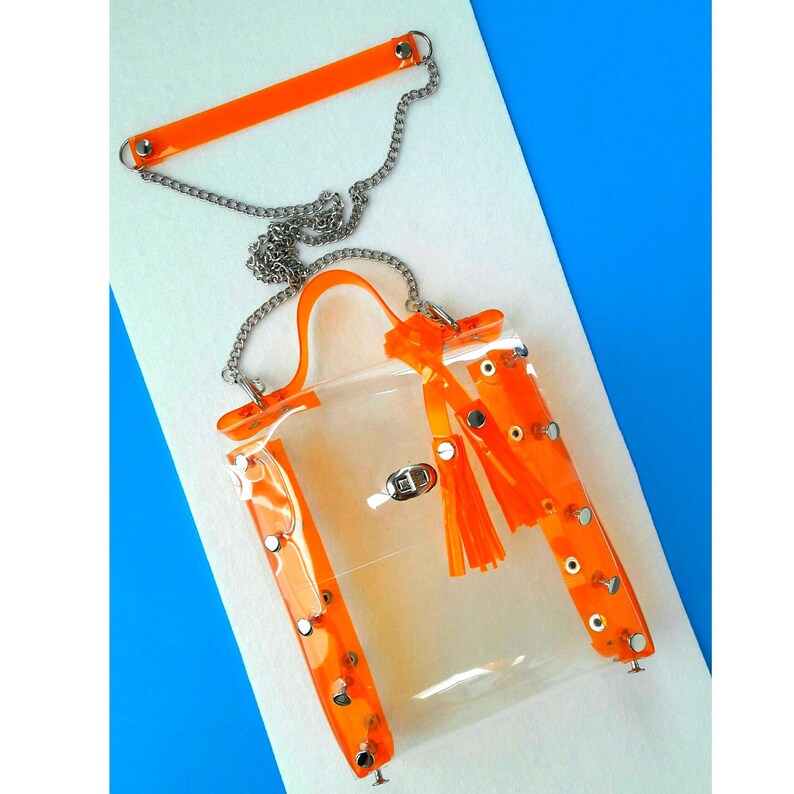 May include: A clear, rectangular handbag with bright orange accents. The bag features a silver chain strap and orange trim with silver rivets. A matching orange handle and tassel add to the design. The bag is set against a white and blue background.