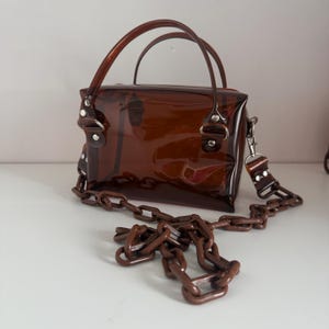 May include: A transparent brown handbag with a rectangular shape. It has two curved handles and a detachable chain strap. The bag features silver-coloured hardware and a glossy finish, suitable for carrying essentials.