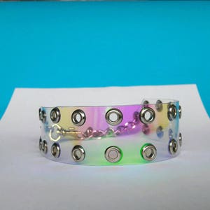 May include: A clear, iridescent choker with silver grommets and a silver chain closure. The choker has a rainbow sheen, reflecting colors of pink, yellow, and green. The accessory is displayed on a white surface against a blue backdrop.