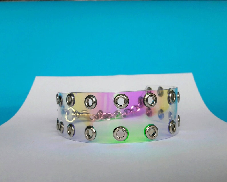 May include: A clear, iridescent choker with silver grommets and a silver chain closure. The choker has a rainbow sheen, reflecting colors of pink, yellow, and green. The accessory is displayed on a white surface against a blue backdrop.