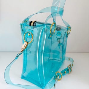 May include: A transparent turquoise handbag with gold-colored hardware. The bag features a top handle and a detachable shoulder strap. The bag's design includes gold-colored rings and a zipper closure. The bag is made of a clear, flexible material.