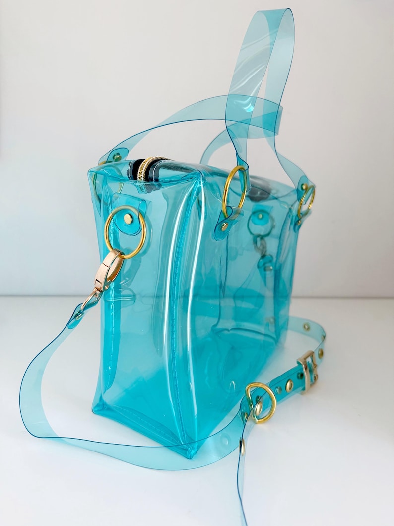 May include: A transparent turquoise handbag with gold-colored hardware. The bag features a top handle and a detachable shoulder strap. The bag's design includes gold-colored rings and a zipper closure. The bag is made of a clear, flexible material.