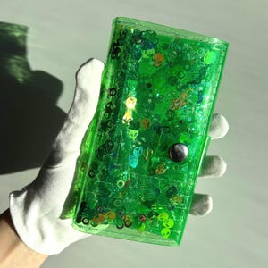 May include: A transparent green wallet filled with glitter and small alien-themed shapes. The wallet has a snap closure and is held by a gloved hand. The glitter and shapes are various colors.