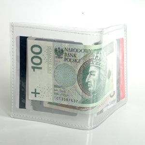 May include: A clear, transparent bifold wallet featuring white stitched edges. The wallet contains a 100 Polish Zloty banknote with the text Narodowy Bank Polski and Wladyslaw II Jagiello, along with several credit cards tucked into the interior slots.
