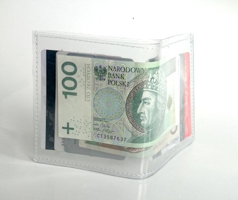 May include: A clear, transparent bifold wallet featuring white stitched edges. The wallet contains a 100 Polish Zloty banknote with the text Narodowy Bank Polski and Wladyslaw II Jagiello, along with several credit cards tucked into the interior slots.