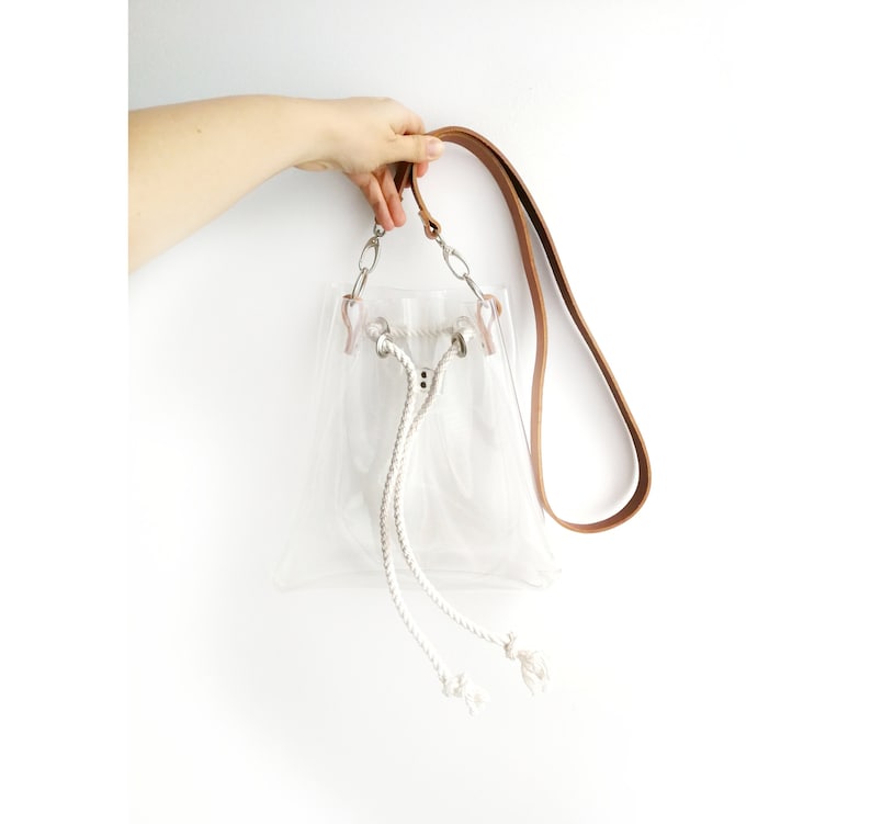 May include: A clear transparent bucket bag featuring a tan leather shoulder strap, silver-toned metal hardware, and a white braided rope drawstring closure with knotted ends. The bag has a structured base and a simple, open-top design.