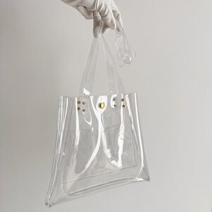 K&ouml;nnte beinhalten: Transparente Tragetasche mit transparenten Griffen und einer kleinen Tasche. Die Tasche wird von einer behandschuhten Hand gehalten. Goldfarbene Nieten befestigen die Griffe an der Tasche. Die Tasche besteht aus transparentem Material.