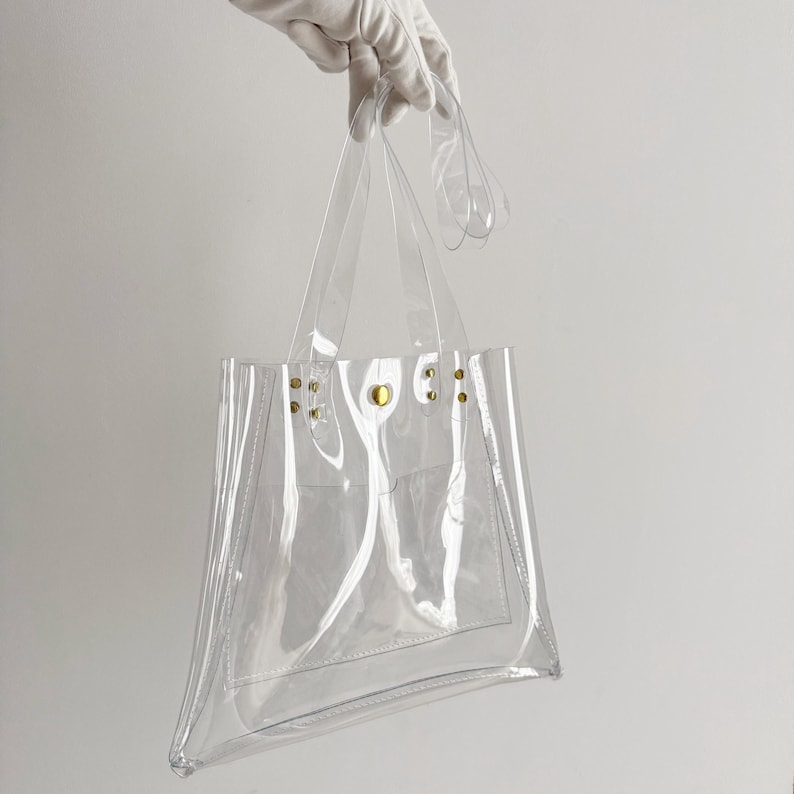 K&ouml;nnte beinhalten: Transparente Tragetasche mit transparenten Griffen und einer kleinen Tasche. Die Tasche wird von einer behandschuhten Hand gehalten. Goldfarbene Nieten befestigen die Griffe an der Tasche. Die Tasche besteht aus transparentem Material.