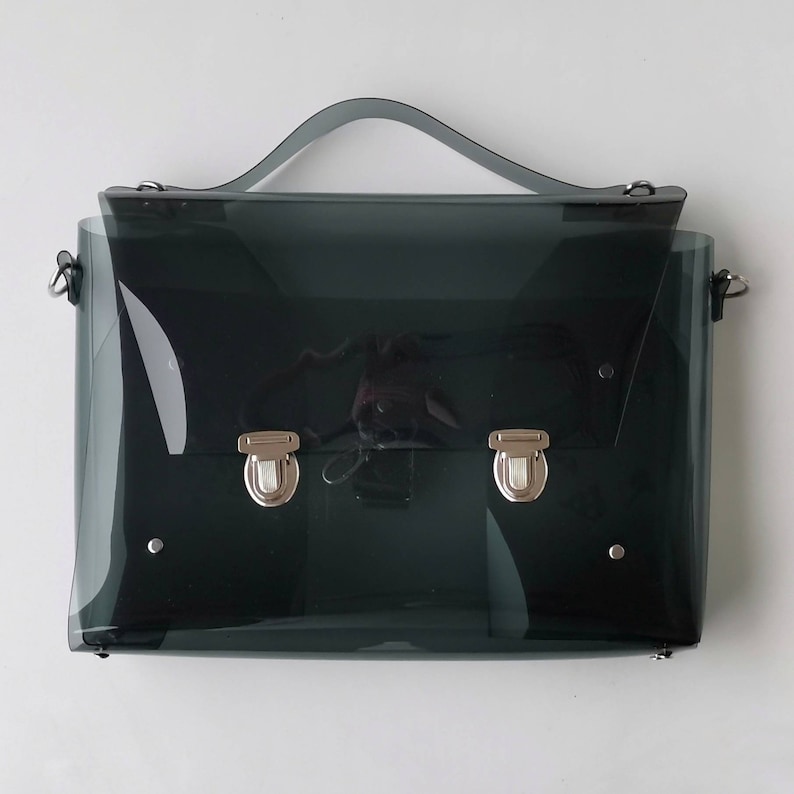 May include: A clear, dark gray plastic briefcase with silver hardware. The bag has a top handle and a shoulder strap.