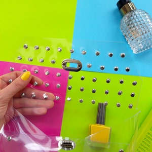 Puede incluir: Una bolsa de pl&aacute;stico transparente con tachuelas plateadas, una botella de esmalte de u&ntilde;as amarillo y una botella de vidrio transparente con tapa plateada. La bolsa est&aacute; sobre un fondo multicolor de rosa, verde y azul.