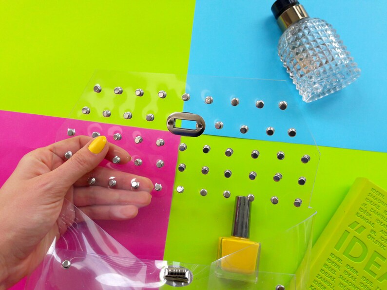 Puede incluir: Una bolsa de pl&aacute;stico transparente con tachuelas plateadas, una botella de esmalte de u&ntilde;as amarillo y una botella de vidrio transparente con tapa plateada. La bolsa est&aacute; sobre un fondo multicolor de rosa, verde y azul.