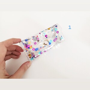 May include: A clear plastic pouch with a silver clasp. The pouch is filled with colorful glitter and sequins, including stars, hearts, and circles.