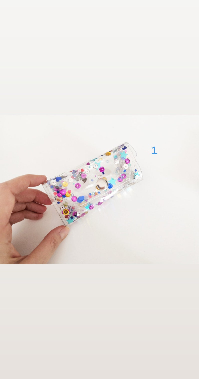 May include: A clear plastic pouch with a silver clasp. The pouch is filled with colorful glitter and sequins, including stars, hearts, and circles.