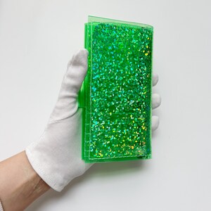May include: A clear, rectangular, green glitter wallet. The wallet is held by a gloved hand. The glitter inside the wallet is a bright green color and reflects light.