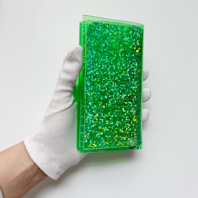 May include: A clear, rectangular, green glitter wallet. The wallet is held by a gloved hand. The glitter inside the wallet is a bright green color and reflects light.