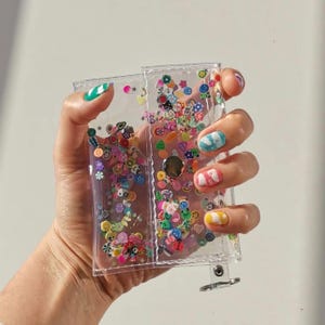 May include: A clear, rectangular, plastic wallet filled with colorful, small, decorative shapes. The wallet has a silver clasp and is held in a hand with painted nails. The shapes include flowers, fruits, and smiley faces.
