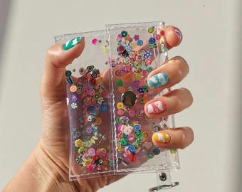 May include: A clear, rectangular, plastic wallet filled with colorful, small, decorative shapes. The wallet has a silver clasp and is held in a hand with painted nails. The shapes include flowers, fruits, and smiley faces.