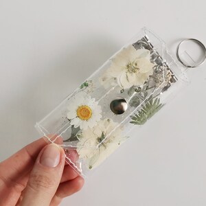 May include: A clear plastic keychain with a silver ring and a button closure. The keychain is filled with pressed white flowers and greenery.