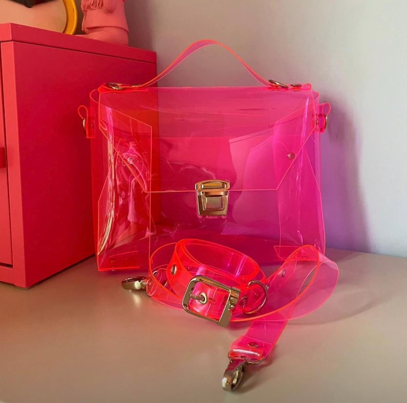 May include: A transparent neon pink handbag with a top handle and a detachable shoulder strap. The bag has a rectangular shape with a gold-colored clasp and hardware. The strap and handle are also neon pink.