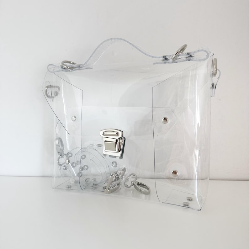 May include: A clear, transparent handbag with a top handle and a shoulder strap. The bag features silver-colored hardware, including a clasp closure, rivets, and rings. The bag is made of a clear, see-through material.