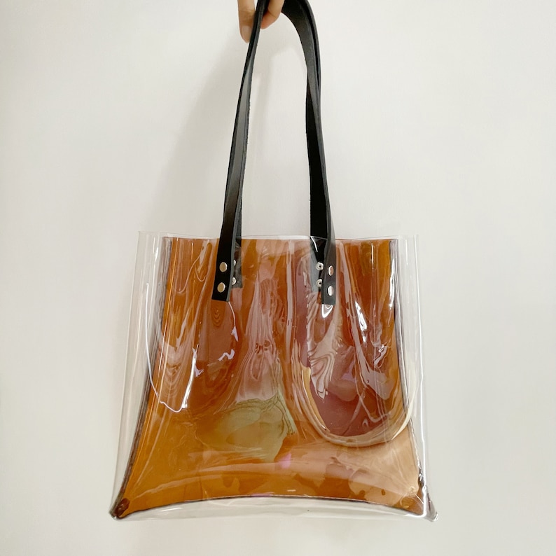 Puede incluir: Bolso tote transparente con interior marr&oacute;n y asas de cuero negro. El bolso es rectangular con un cuerpo transparente, lo que permite ver el contenido. Las asas est&aacute;n sujetas con remaches plateados.