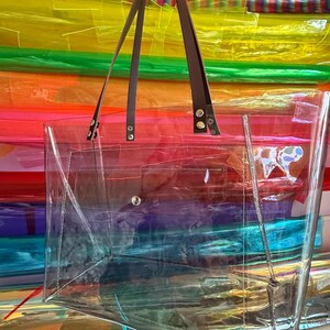 Puede incluir: Un bolso tote rectangular transparente con dos correas de hombro marrones sujetas con remaches de metal plateado. El bolso tiene un cuerpo transparente con costuras visibles en los bordes y un cierre de bot&oacute;n a presi&oacute;n plateado en el centro.