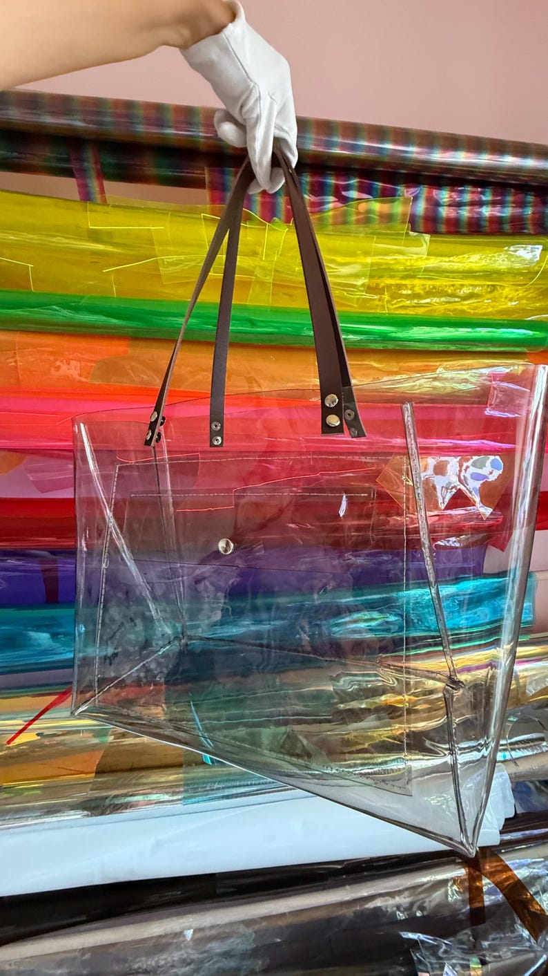 Puede incluir: Un bolso tote rectangular transparente con dos correas de hombro marrones sujetas con remaches de metal plateado. El bolso tiene un cuerpo transparente con costuras visibles en los bordes y un cierre de bot&oacute;n a presi&oacute;n plateado en el centro.