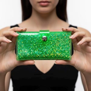 May include: A rectangular, transparent green clutch filled with green and yellow glitter. The clutch has a silver snap closure and is held by a person. The clutch is a vibrant accessory.