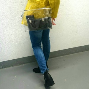 May include: A clear plastic bag with a black leather wallet inside. The bag is worn over the shoulder of a person wearing a yellow jacket and blue jeans.