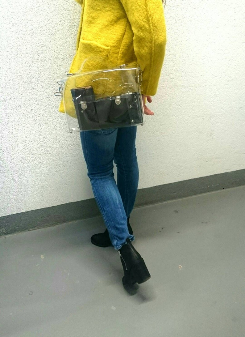 May include: A clear plastic bag with a black leather wallet inside. The bag is worn over the shoulder of a person wearing a yellow jacket and blue jeans.