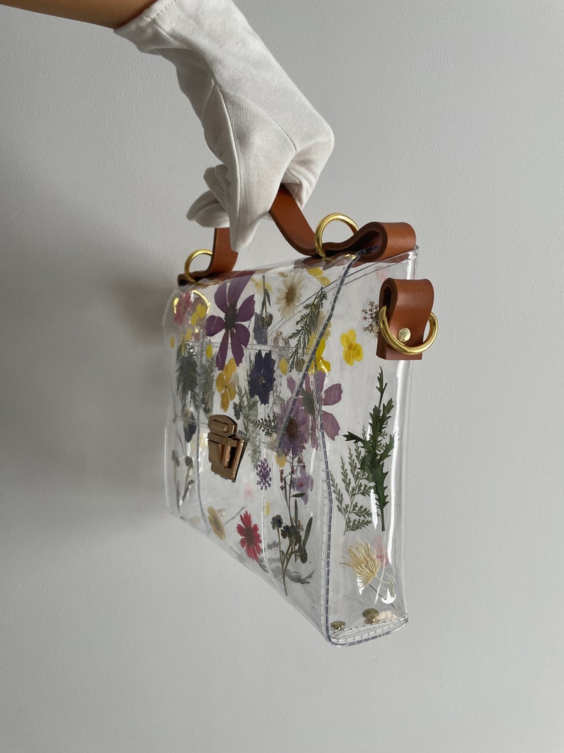 May include: Clear plastic handbag with a brown leather strap and a gold clasp. The bag is decorated with pressed flowers.