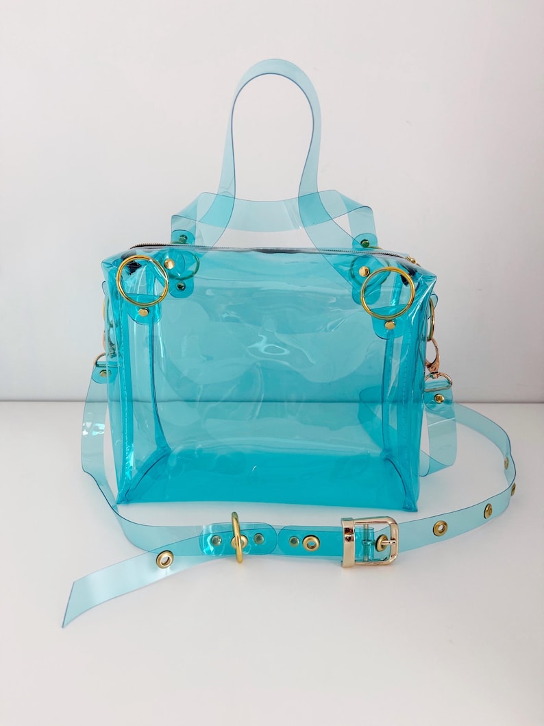 May include: A transparent, turquoise handbag with a top handle and a detachable shoulder strap. The bag features gold-colored hardware, including rings and a buckle. The clear design allows visibility of the contents.