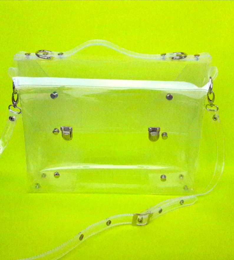 May include: A clear plastic briefcase with a silver handle and silver buckles. The bag has a long adjustable strap.